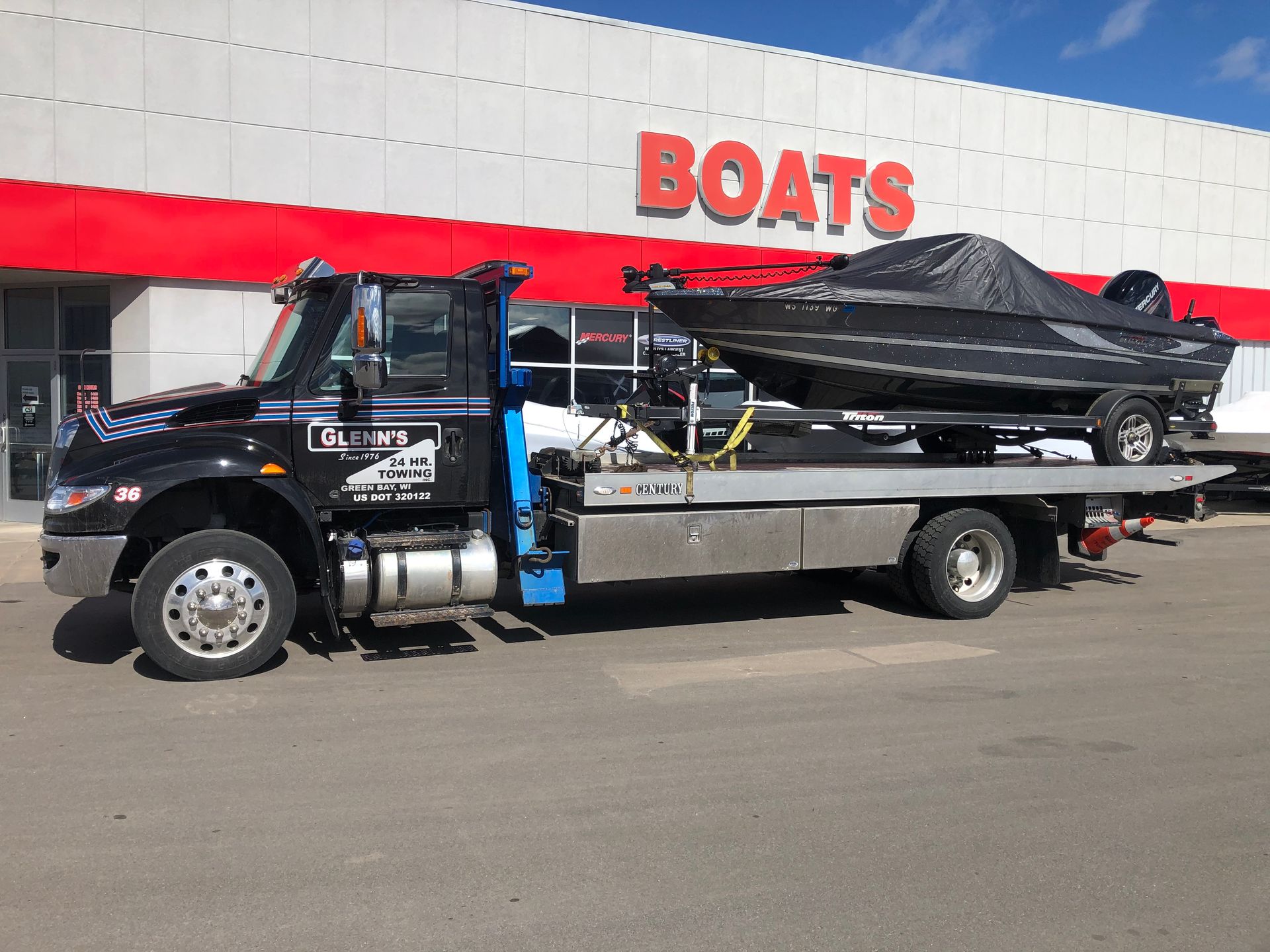 Black tow truck hauling a covered boat in front of a store with 