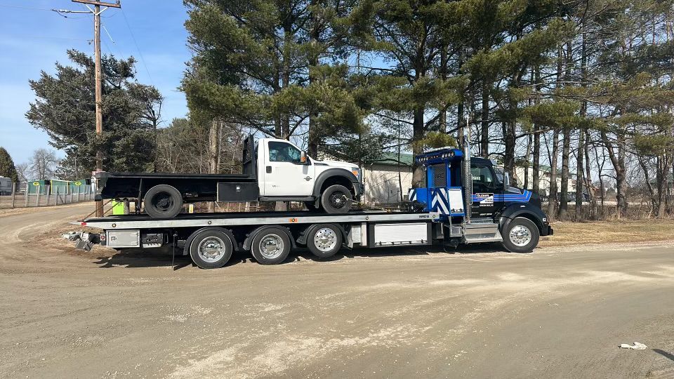 A flatbed tow truck carries a white truck on a paved road with trees in the background.