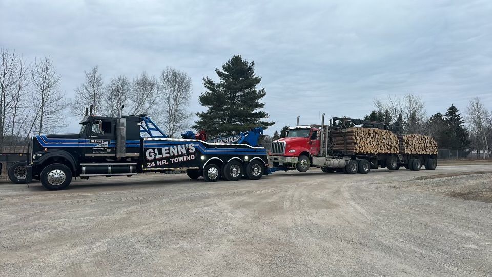 Tow truck towing a red semi-truck hauling logs on a gravel lot under a cloudy sky.