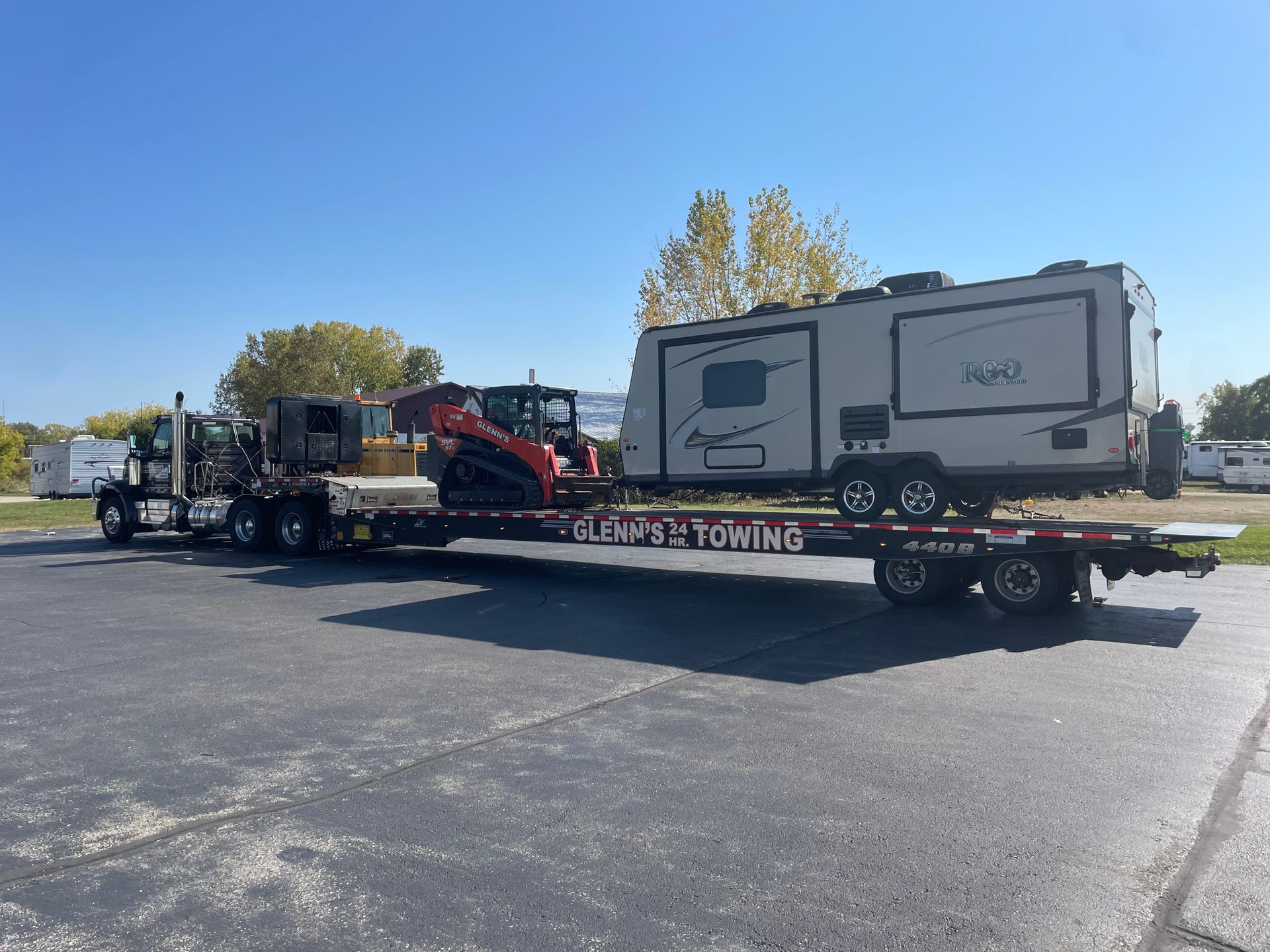 A semi-truck with a flatbed trailer transporting a camper and an orange skid steer.