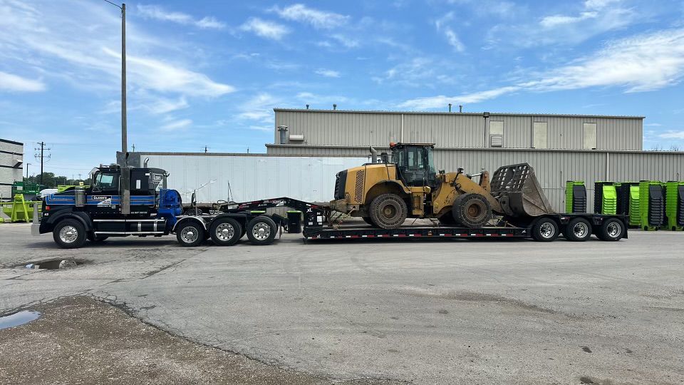 A blue semi-truck hauling a yellow Caterpillar loader on a trailer on a paved lot.