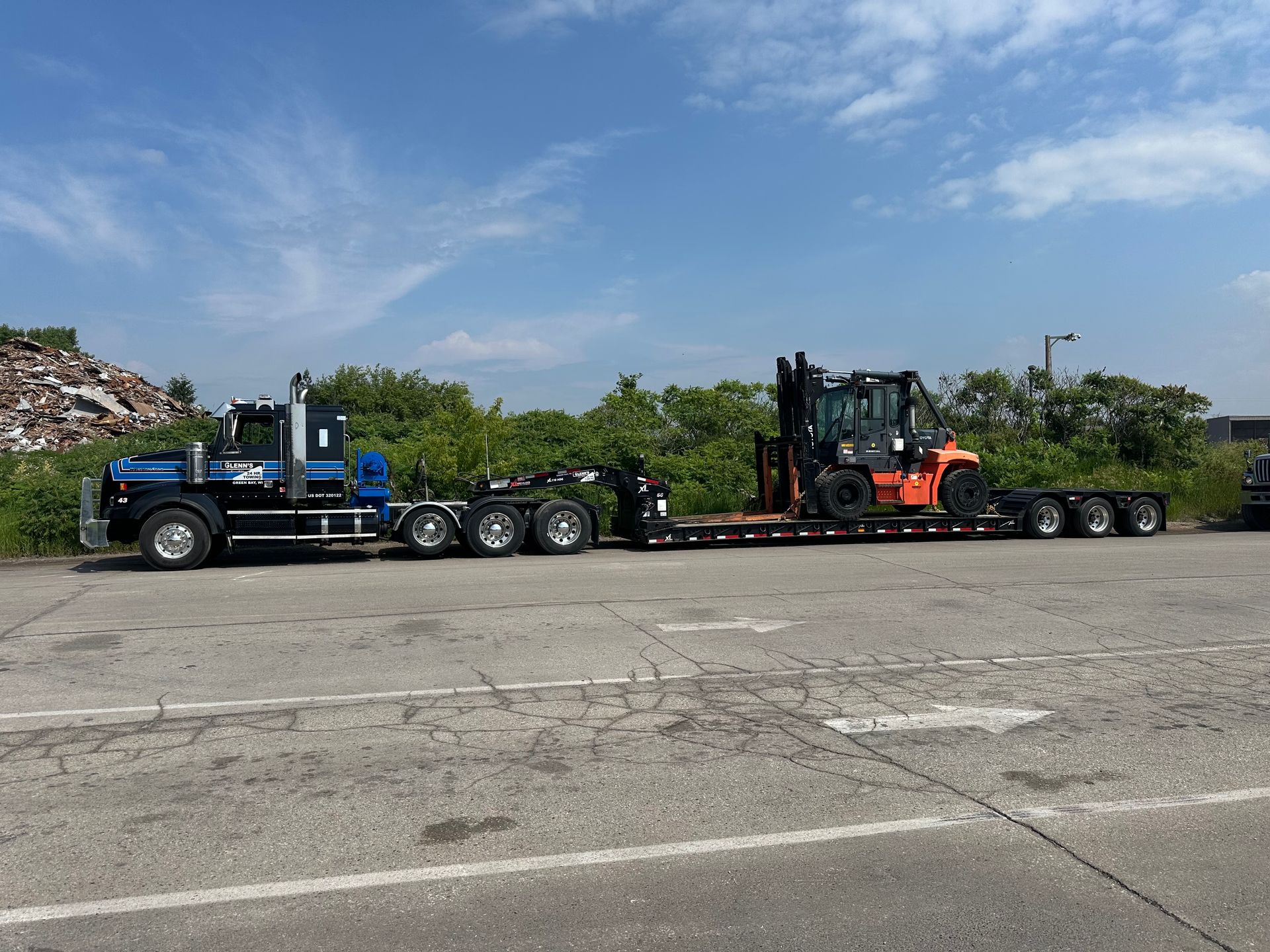 Semi-truck hauling a forklift on a flatbed trailer, parked on asphalt; blue sky overhead.
