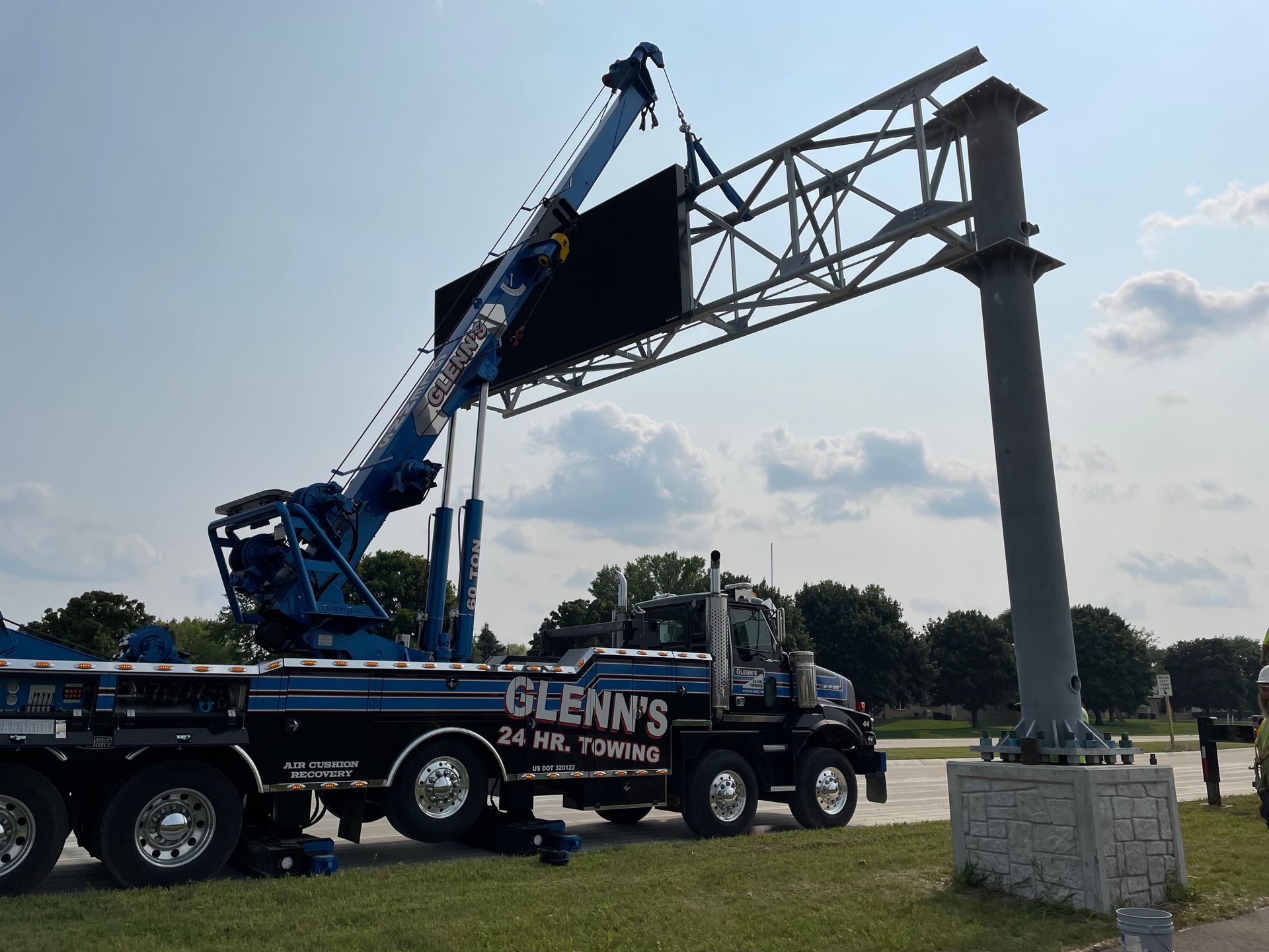 A large crane lifts a highway sign panel from a support column. The crane is attached to a tow truck.