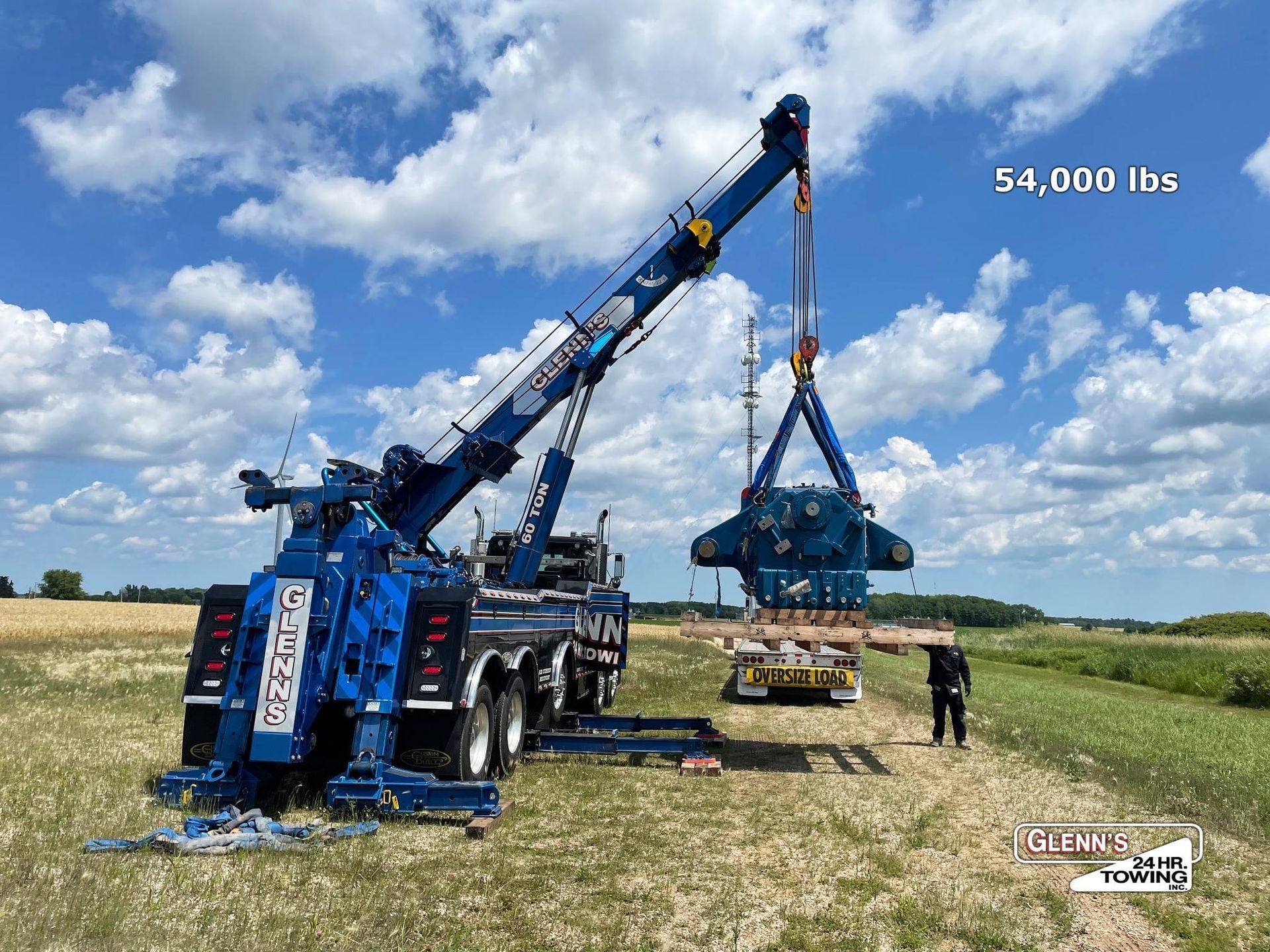 A large blue tow truck lifts a heavy, dark blue object with a crane in a field under a cloudy sky.