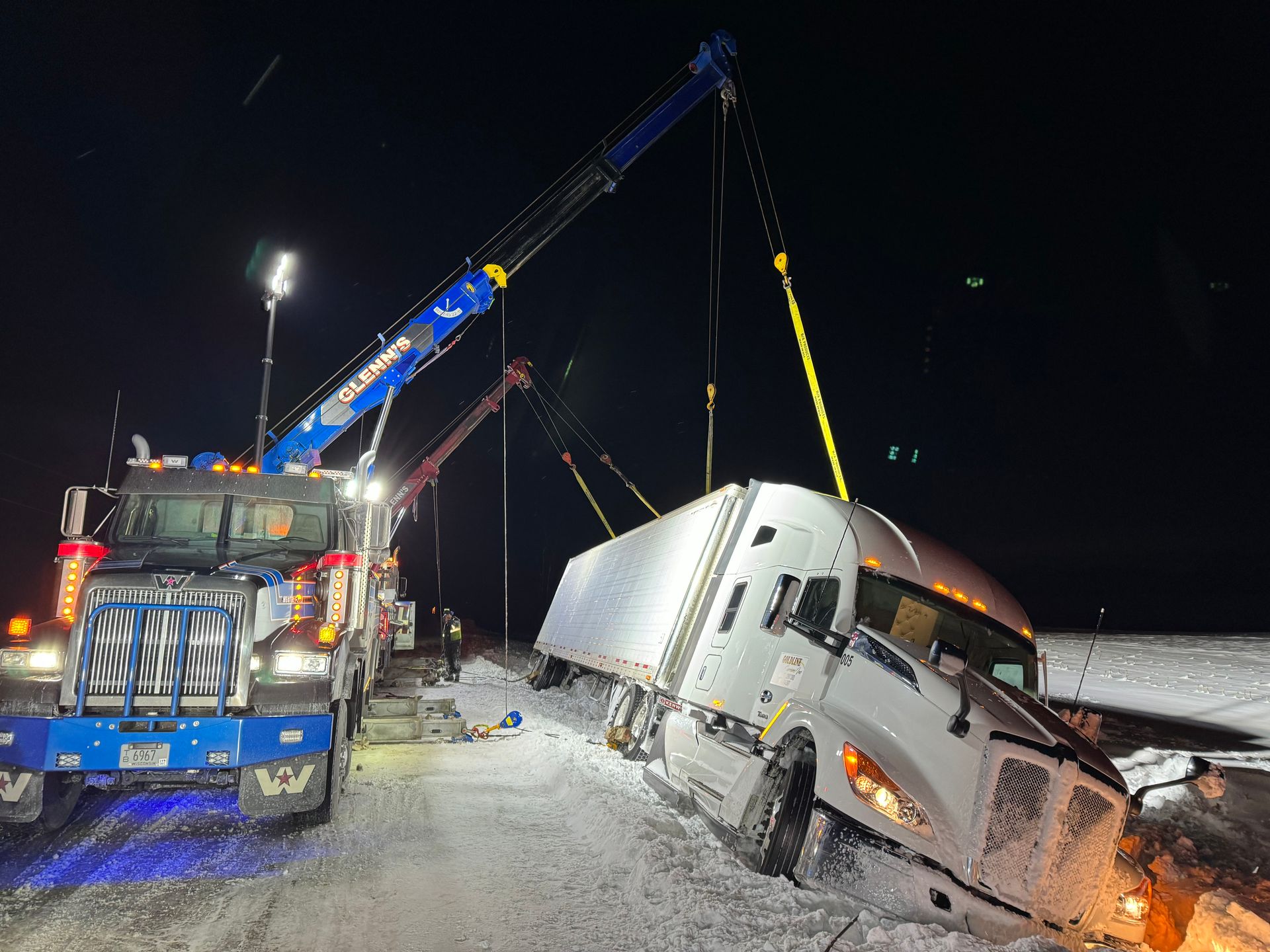 Tow trucks lifting a semi-truck out of a snow-covered ditch at night.