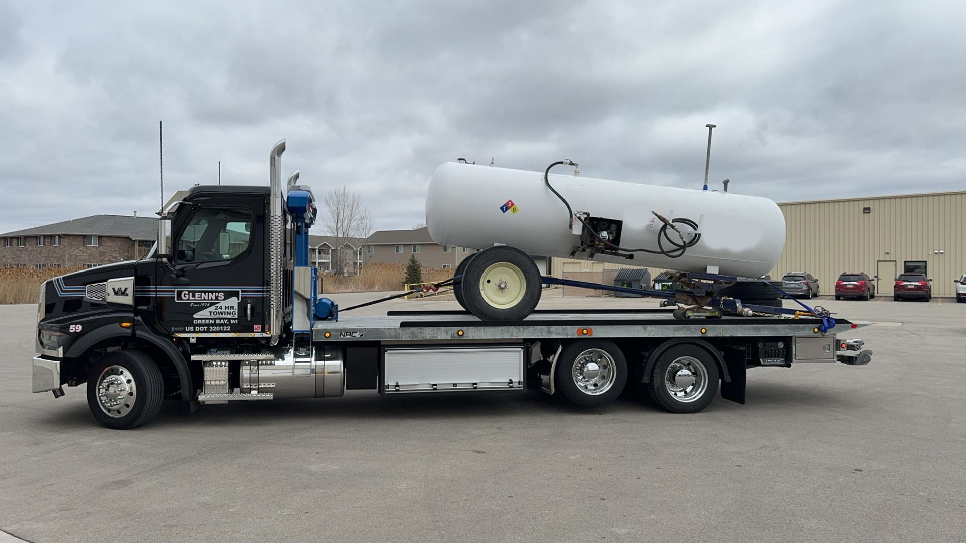 Black flatbed truck carrying a large white tank on wheels, parked outside a building.
