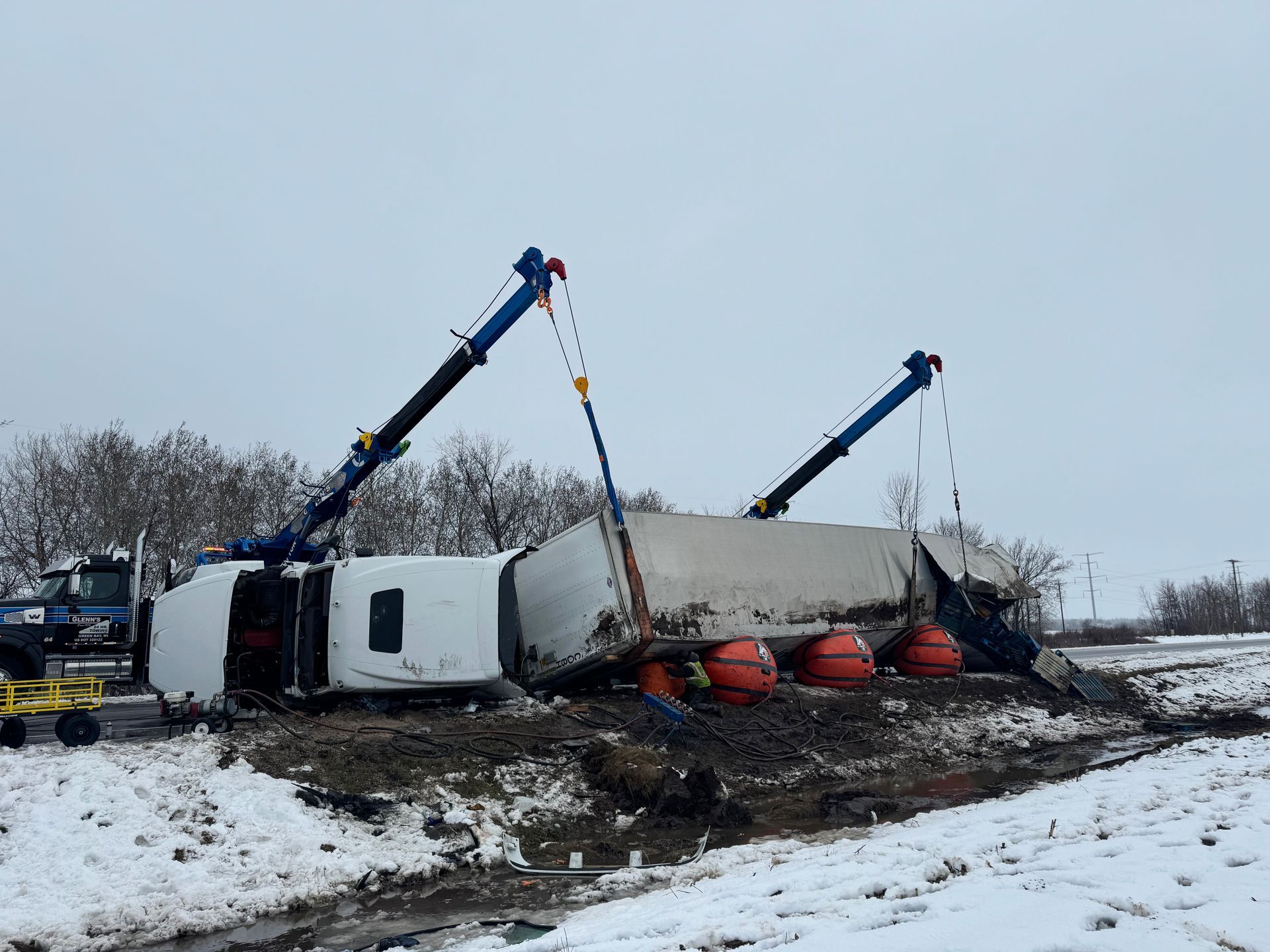 Truck overturned in snow, being lifted by two cranes. White truck, blue cranes, muddy ground.