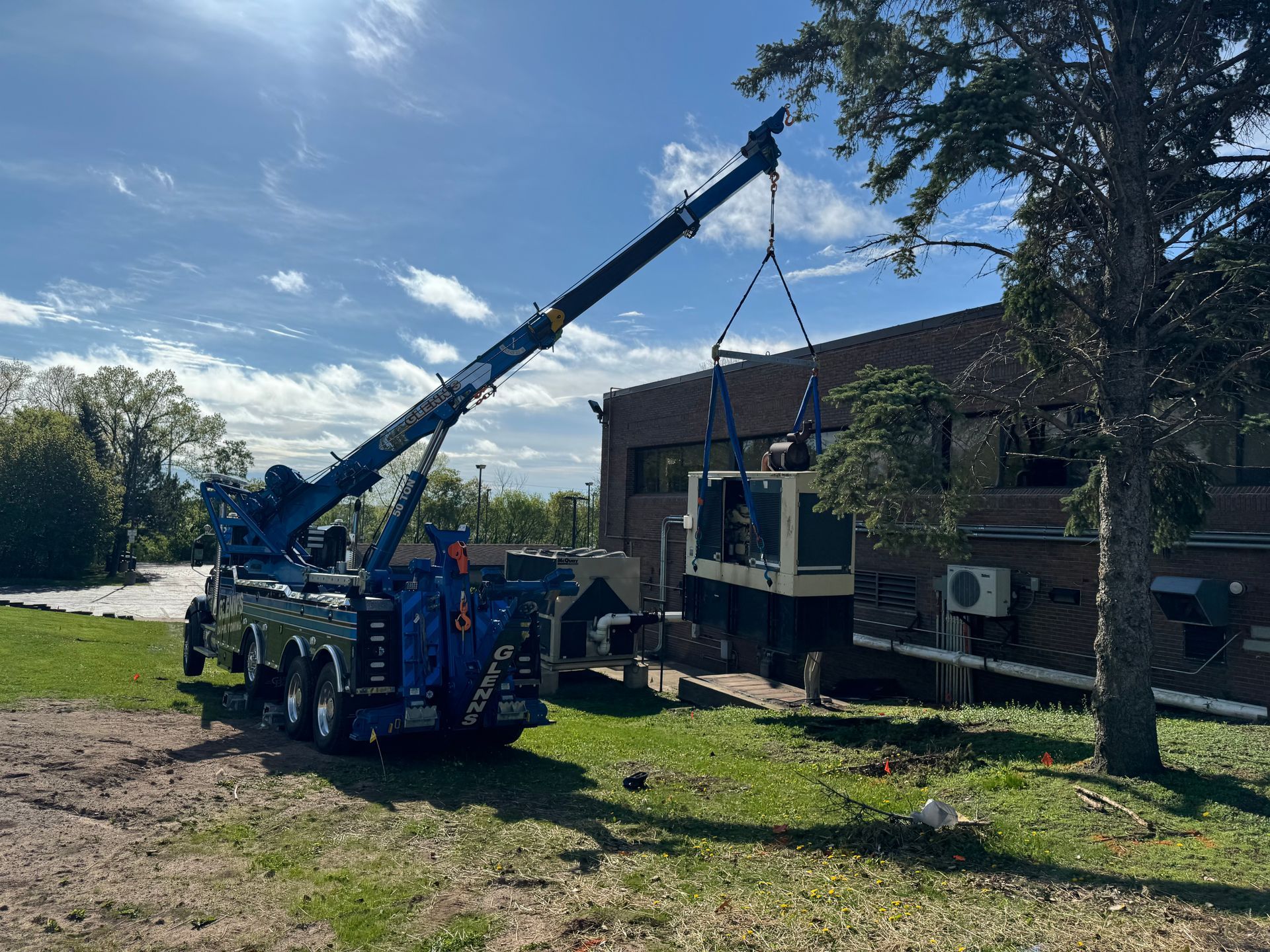 A blue crane lifts a generator near a brick building and a tree on a sunny day.