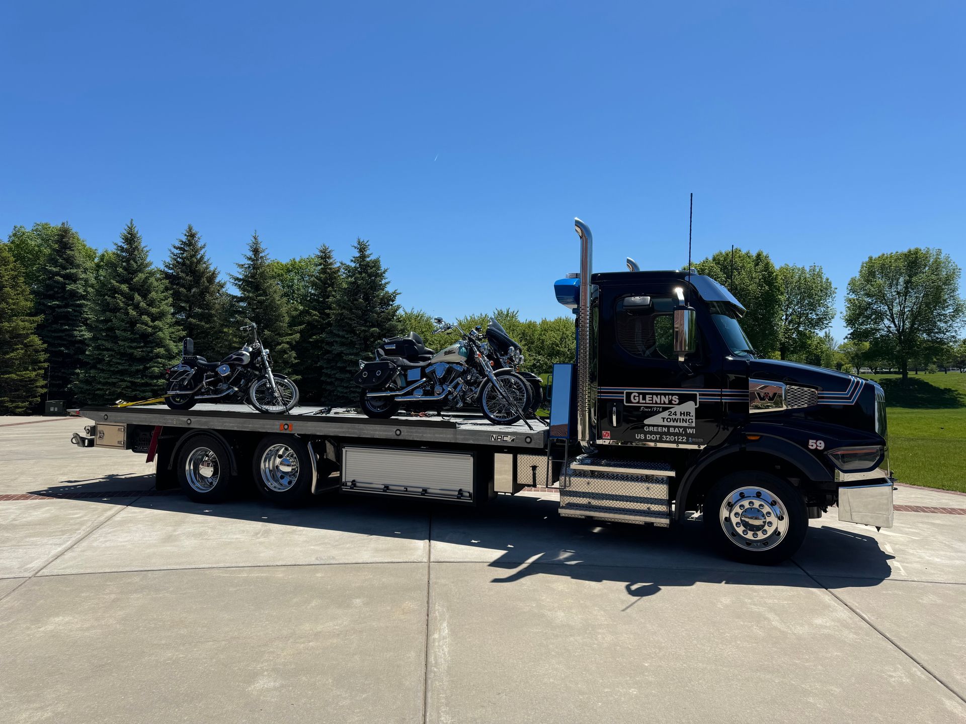Black semi-truck carrying three motorcycles on a flatbed, parked outdoors on a sunny day.