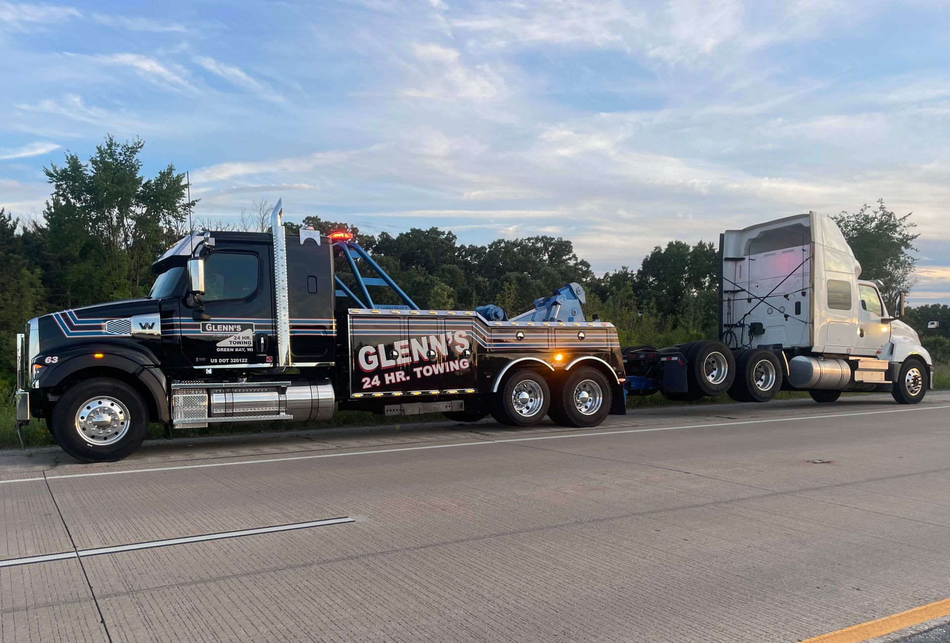 Tow truck towing a semi-truck along a roadside. The tow truck is black with 