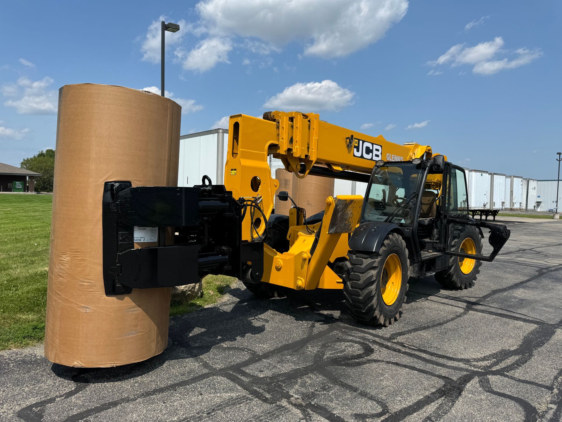 Yellow JCB telehandler lifting a large roll of brown paper on asphalt. Sunny, outdoor setting.