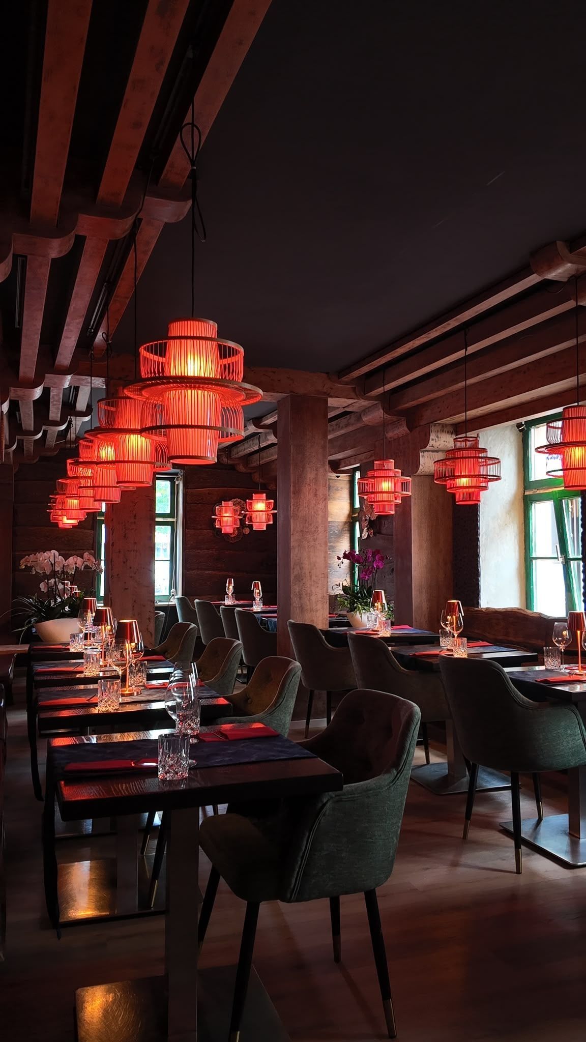 Das Restaurantinterieur ist schwach beleuchtet, mit roten Laternen, dunklen Holztischen und Stühlen in der Nähe großer Fenster.