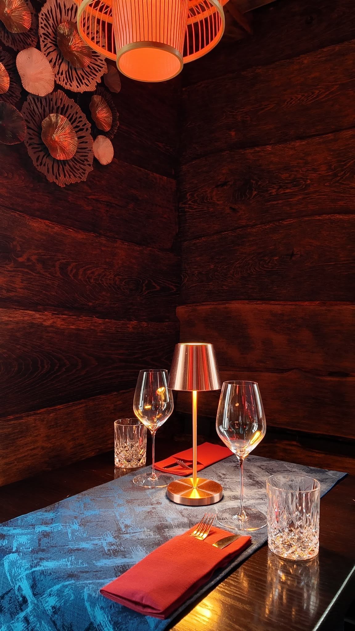 Ein mit Kerzen beleuchteter Restauranttisch mit Weingläsern, Wassergläsern und einer roten Speisekarte vor einer dunklen Holzwand
