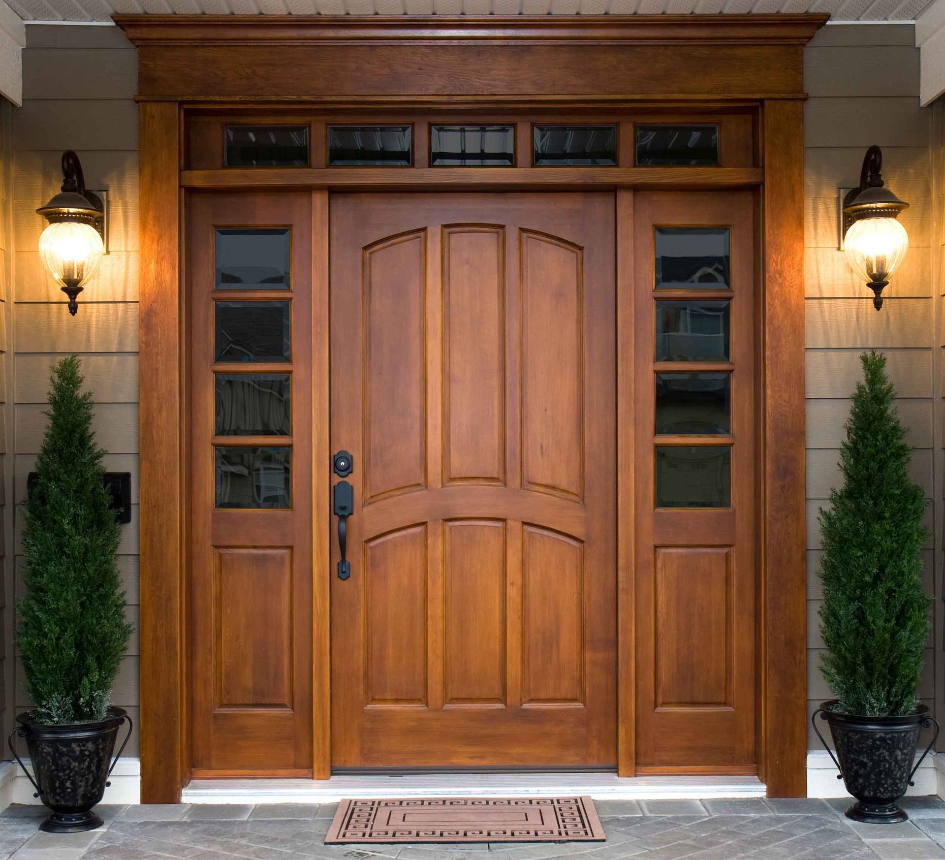 Wooden front door with sidelights and transom, flanked by potted trees and sconce lights.