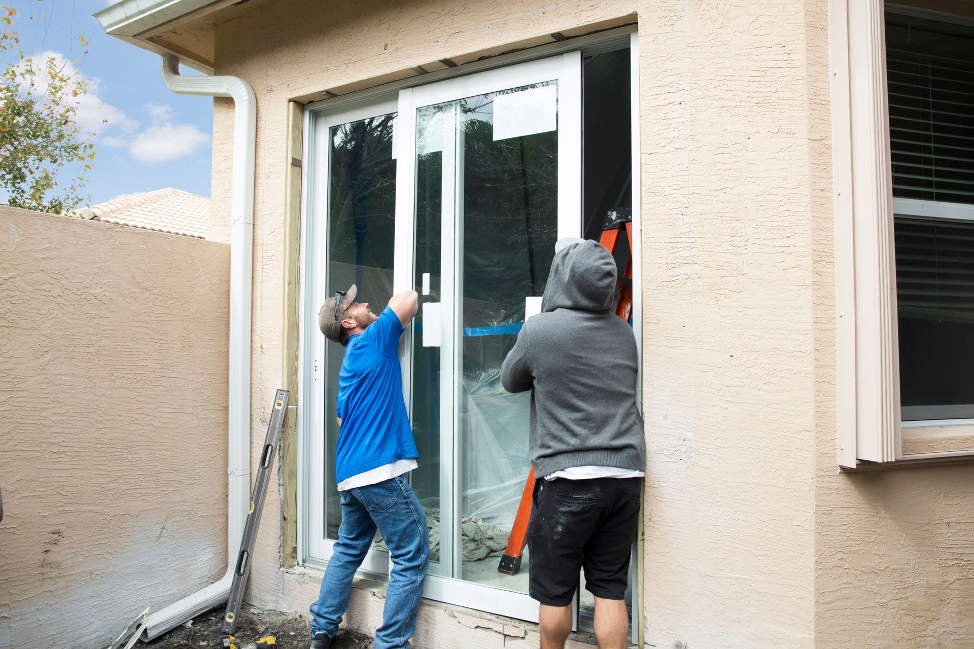 Two people install a sliding glass door in a building. One wears blue, the other a grey hoodie.