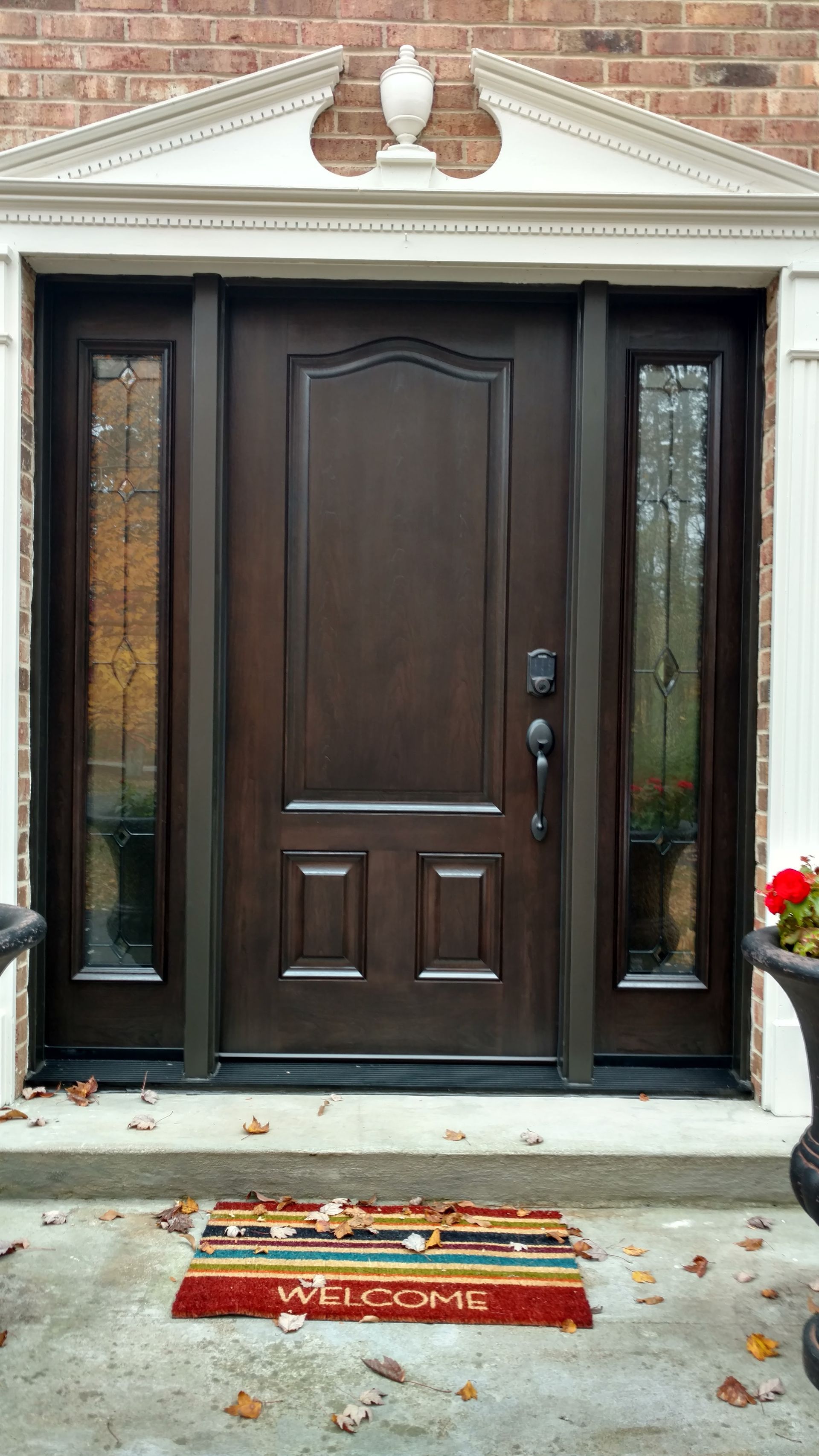 Brown front door with sidelights, welcome mat, beneath a brick facade.