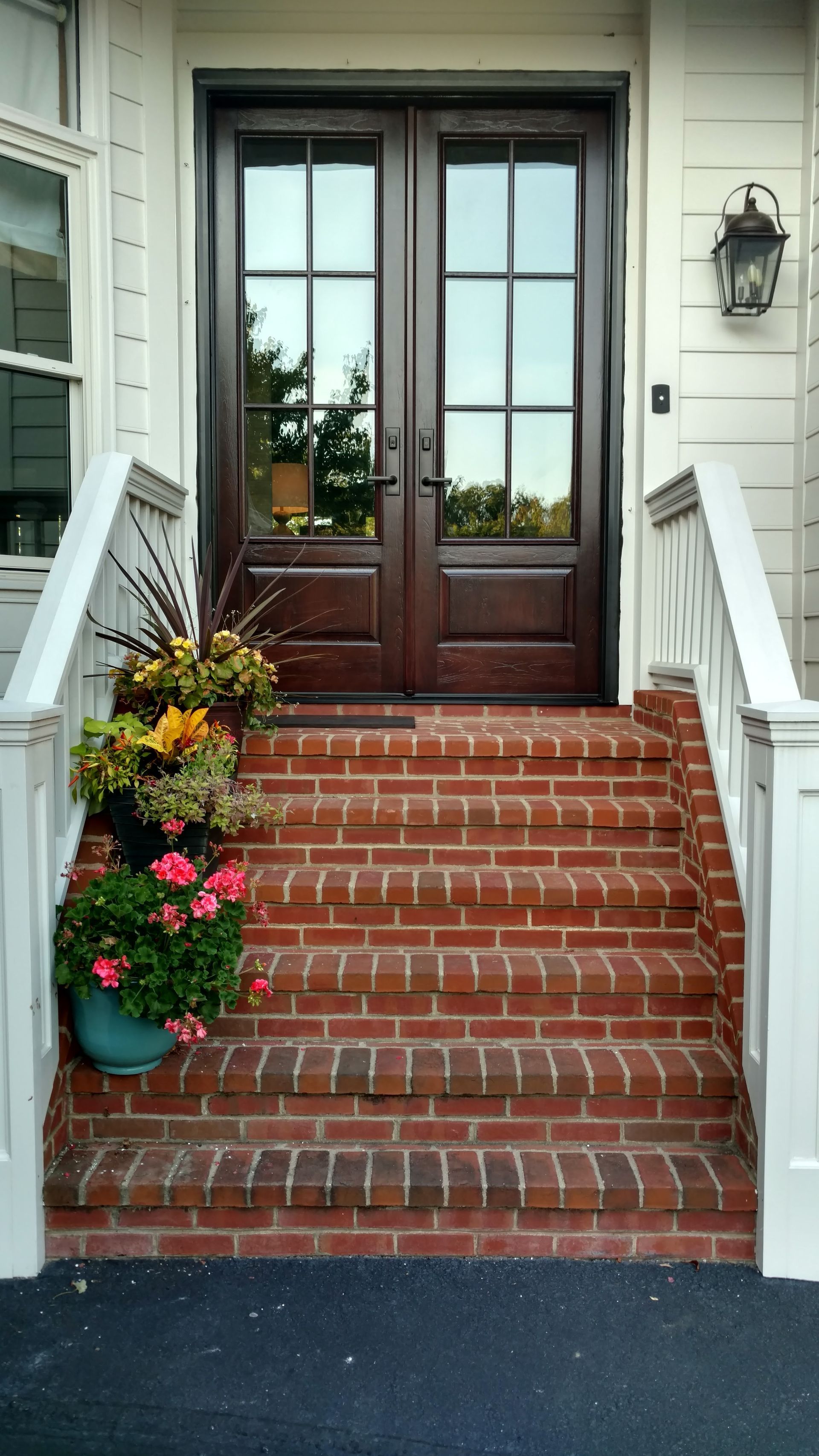 Brick steps lead to brown double doors. White railings flank the steps. Planters with flowers.