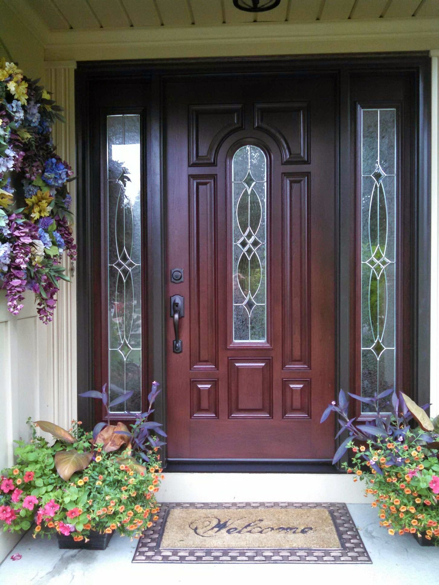 Brown front door with decorative glass panels, flanked by planters with flowers and a welcome mat.