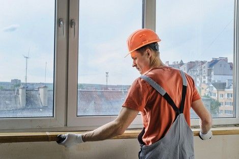 Construction worker installing windowsill, wearing orange hard hat and overalls, working near window, city view.