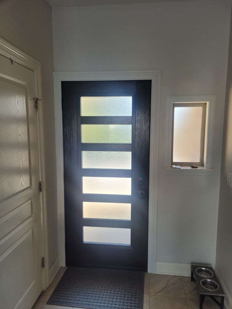 Black front door with frosted glass panels, next to a small window and a white door in an entryway.