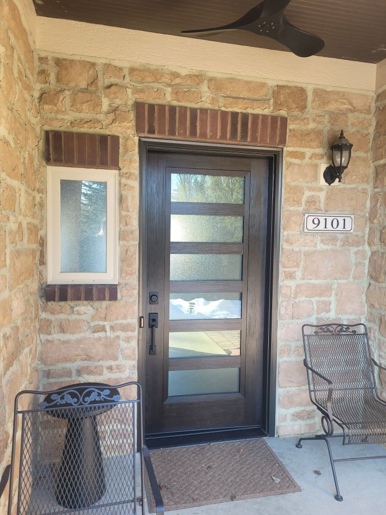 Brown front door with glass panes, brick accents, and stone facade; porch with chair and street number