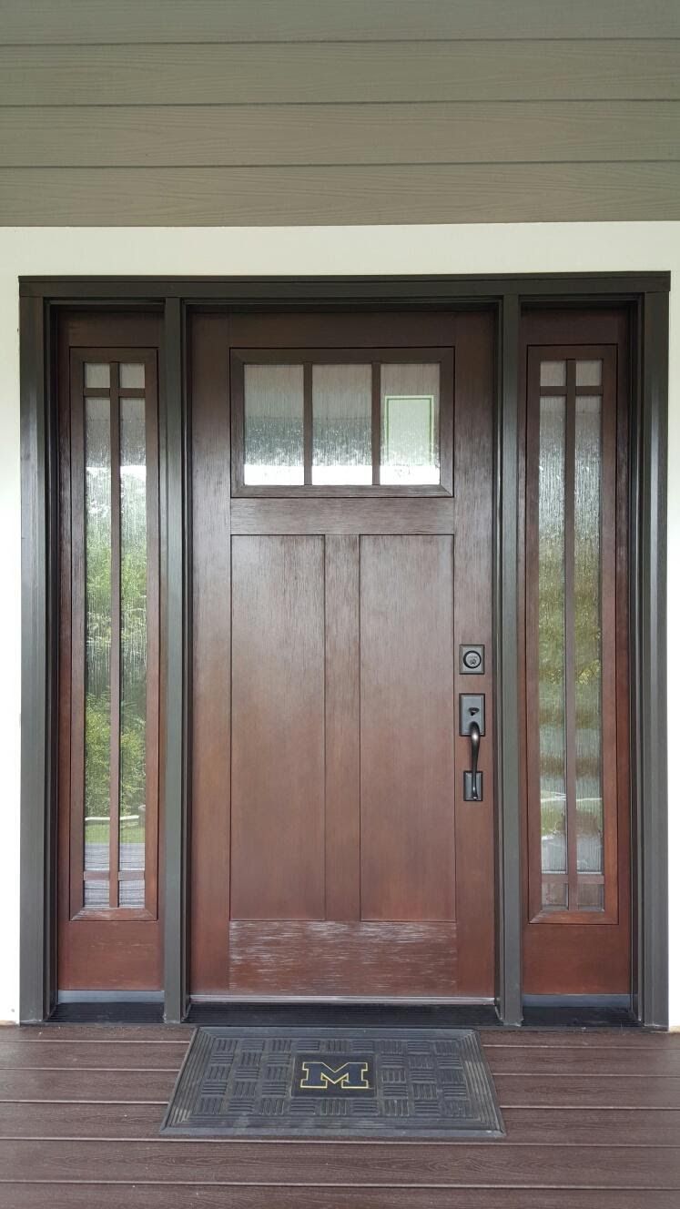 Brown front door with sidelights, under a porch, with a doormat.