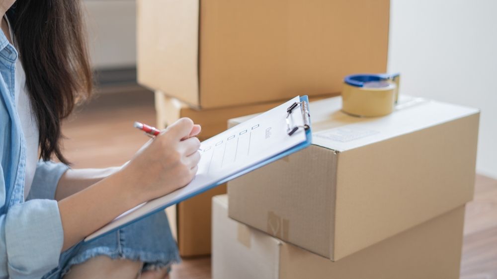 A Woman is Sitting in Front of a Pile of Cardboard Boxes and Writing on a Clipboard — Wide Bay Removals In Dundowran, QLD