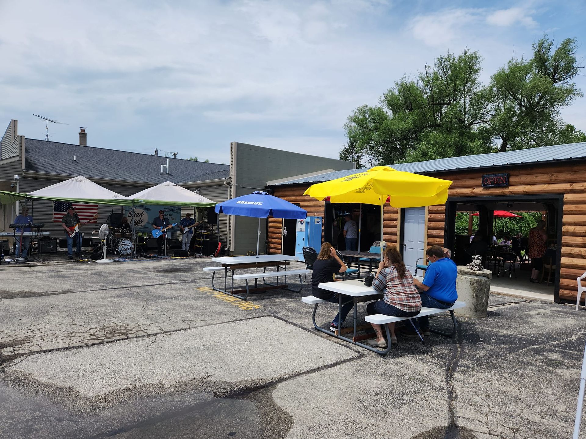 A group of people are sitting at picnic tables under umbrellas in a parking lot.