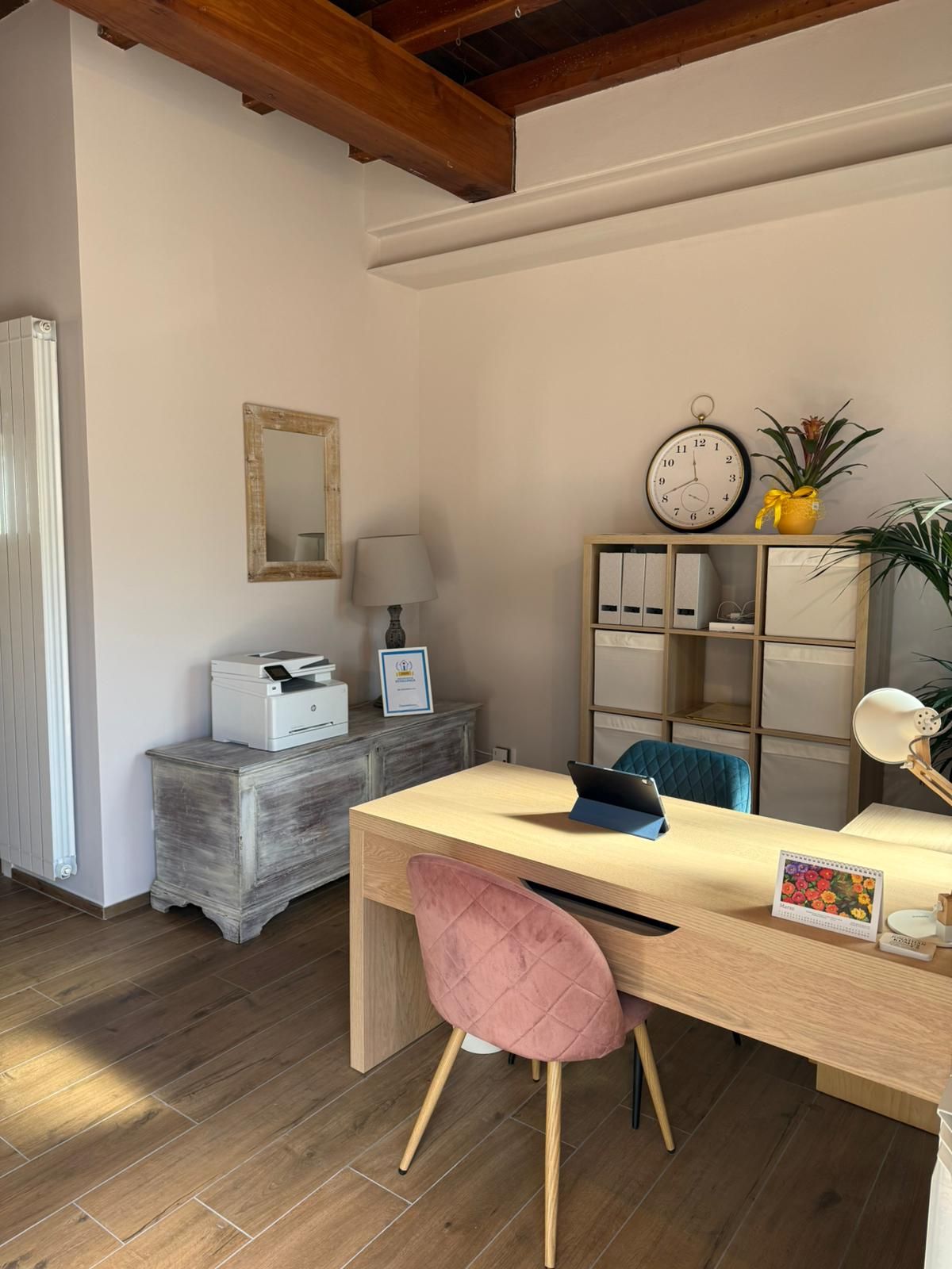 Accogliente ufficio domestico con scrivania in legno, sedia rosa, mensole, lampada, orologio e quadri in una stanza luminosa.