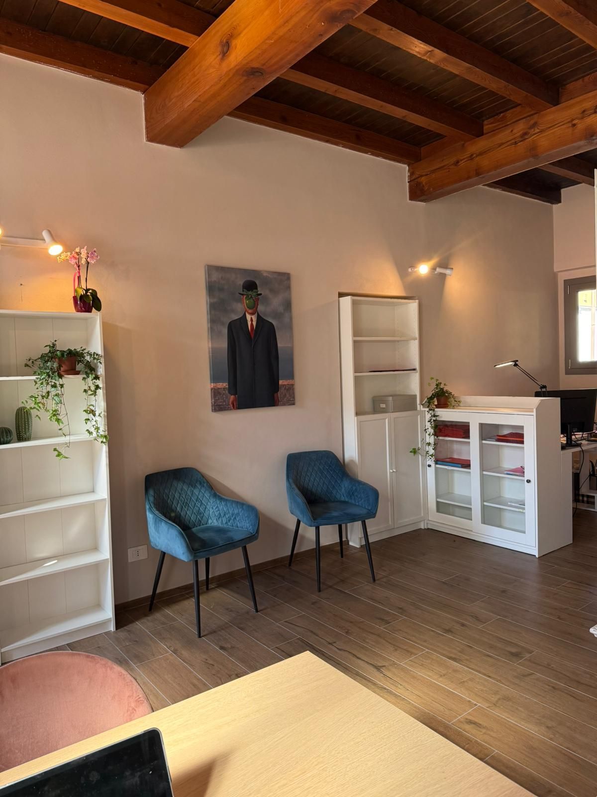 Una stanza accogliente con travi a vista sul soffitto, due sedie blu, mensole bianche e un angolo illuminato dalla finestra.