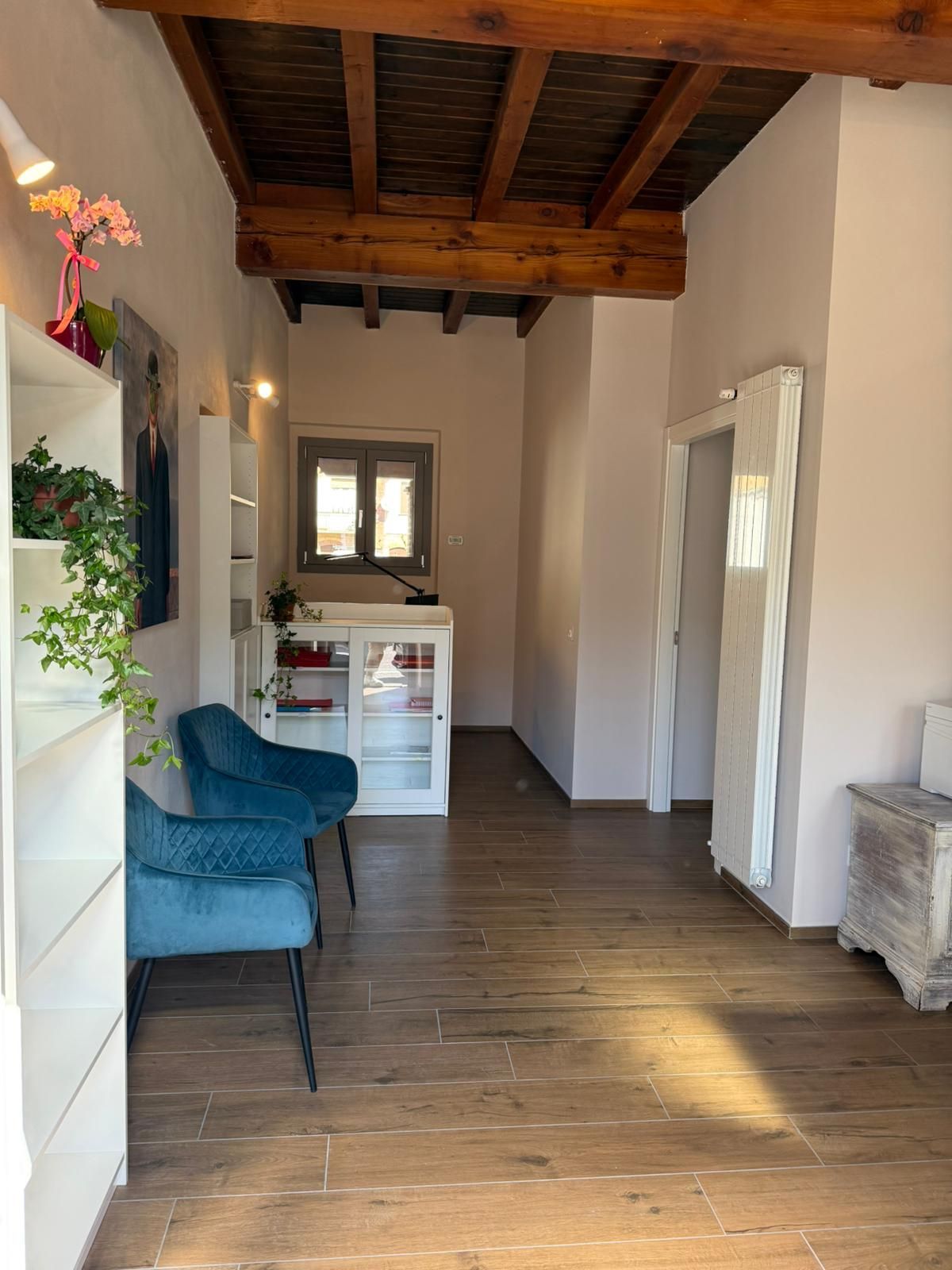 Stretto corridoio con soffitto in legno, sedia blu, piccola scrivania e una finestra in fondo.