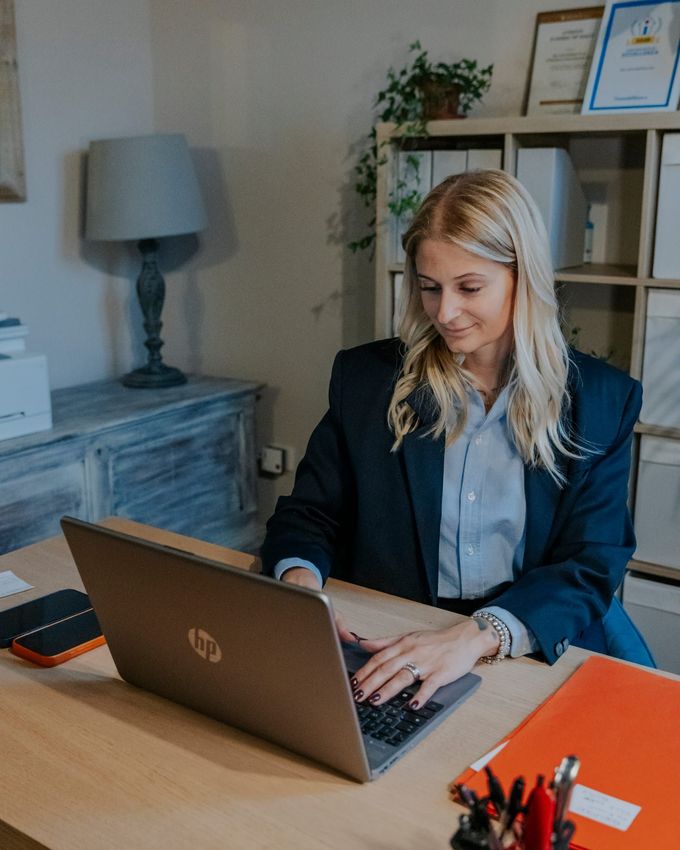 Una donna lavora al computer portatile a una scrivania nel suo ufficio domestico, con una lampada, una libreria e dei certificati incorniciati alle sue spalle.