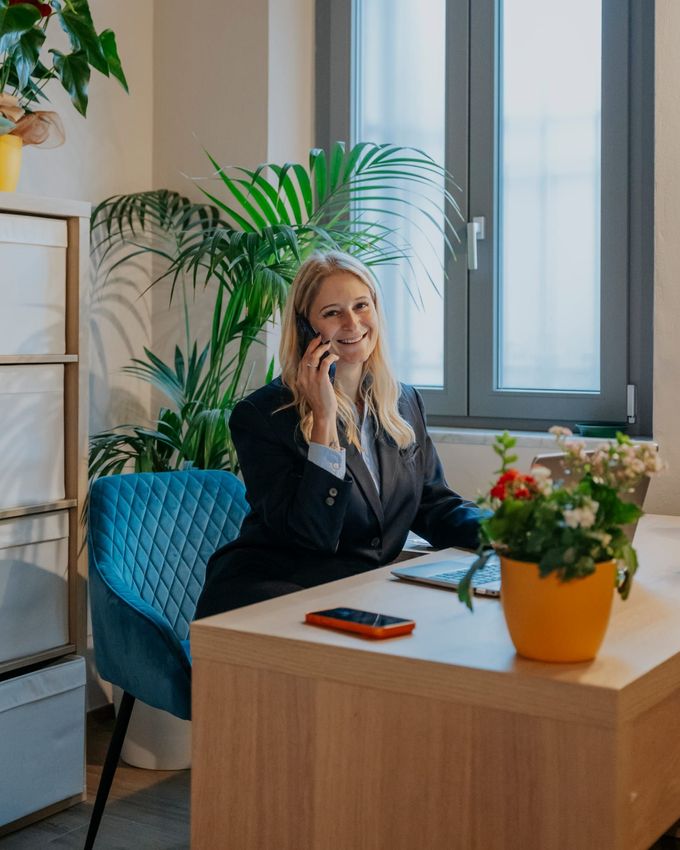Una donna sorride seduta alla sua scrivania, mentre parla al telefono, in un ufficio luminoso con piante e fiori.