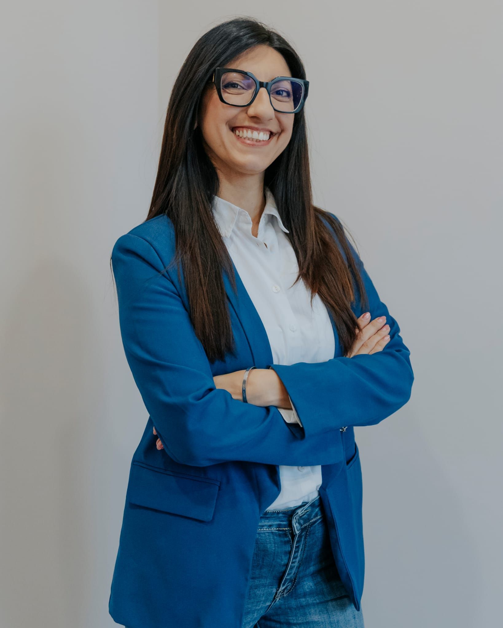 Persona sorridente con blazer blu e camicia bianca, in piedi con le braccia incrociate su uno sfondo grigio uniforme.