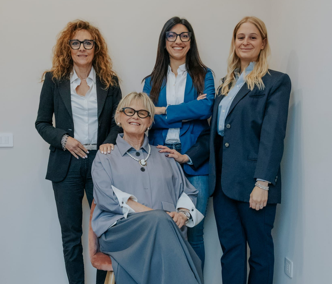 Quattro donne in abiti da lavoro posano insieme, una seduta in primo piano, contro una semplice parete interna.