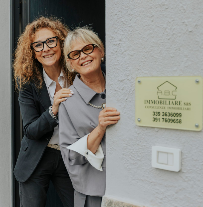 Due persone sorridenti in abiti da lavoro in piedi accanto all'insegna e all'ingresso di un'agenzia immobiliare.