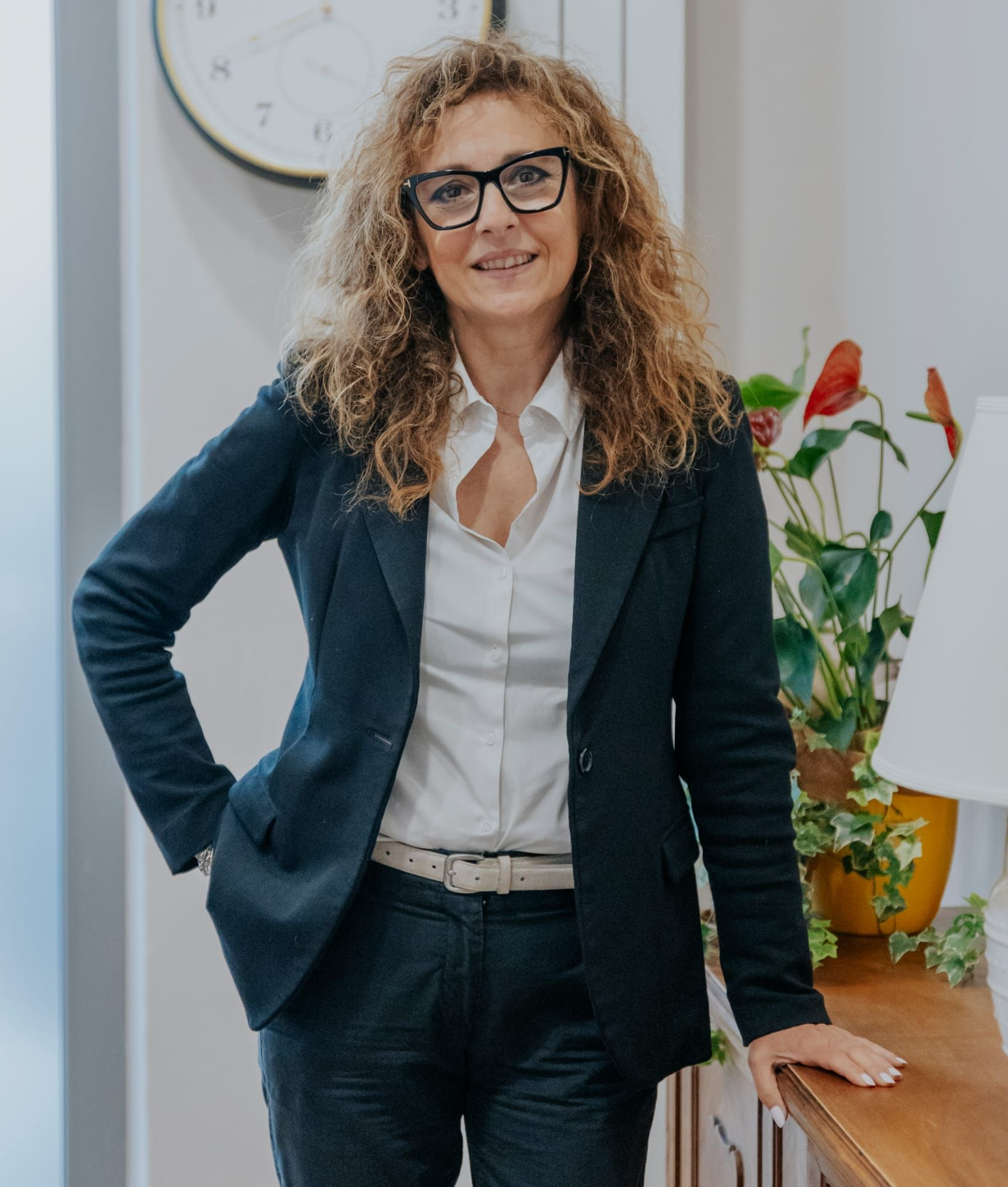 Una donna con un blazer blu e occhiali è in piedi accanto a un tavolo con delle piante, sorridente, in un ambiente interno.