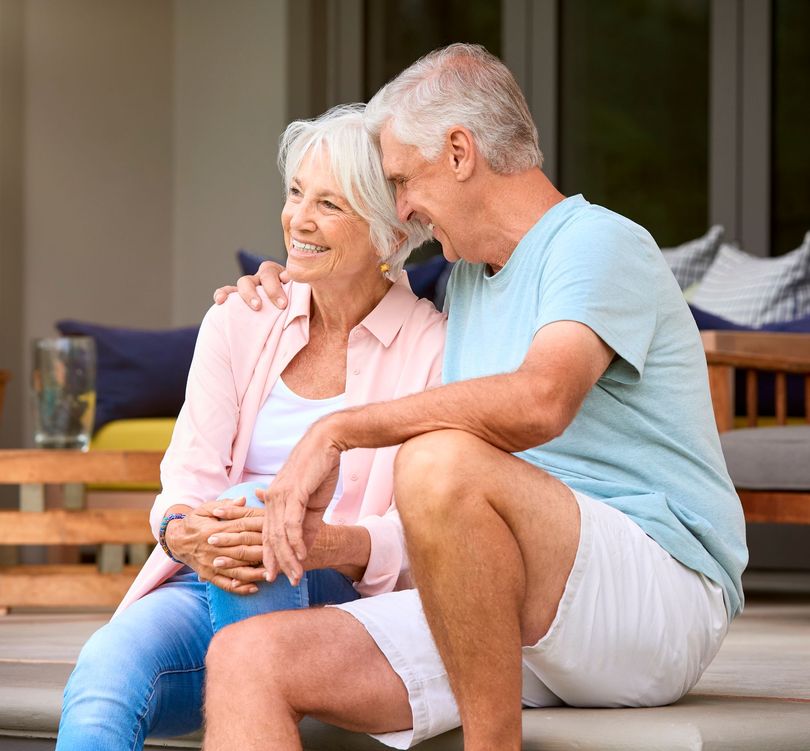 Elderly couple smiling and embracing outdoors on a porch.