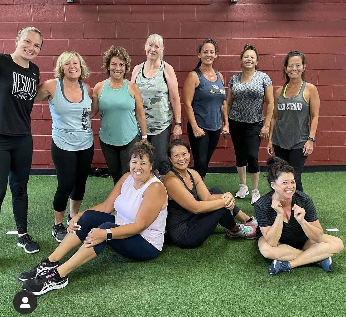 Group of women in athletic wear, smiling, posing on green turf inside a gym.