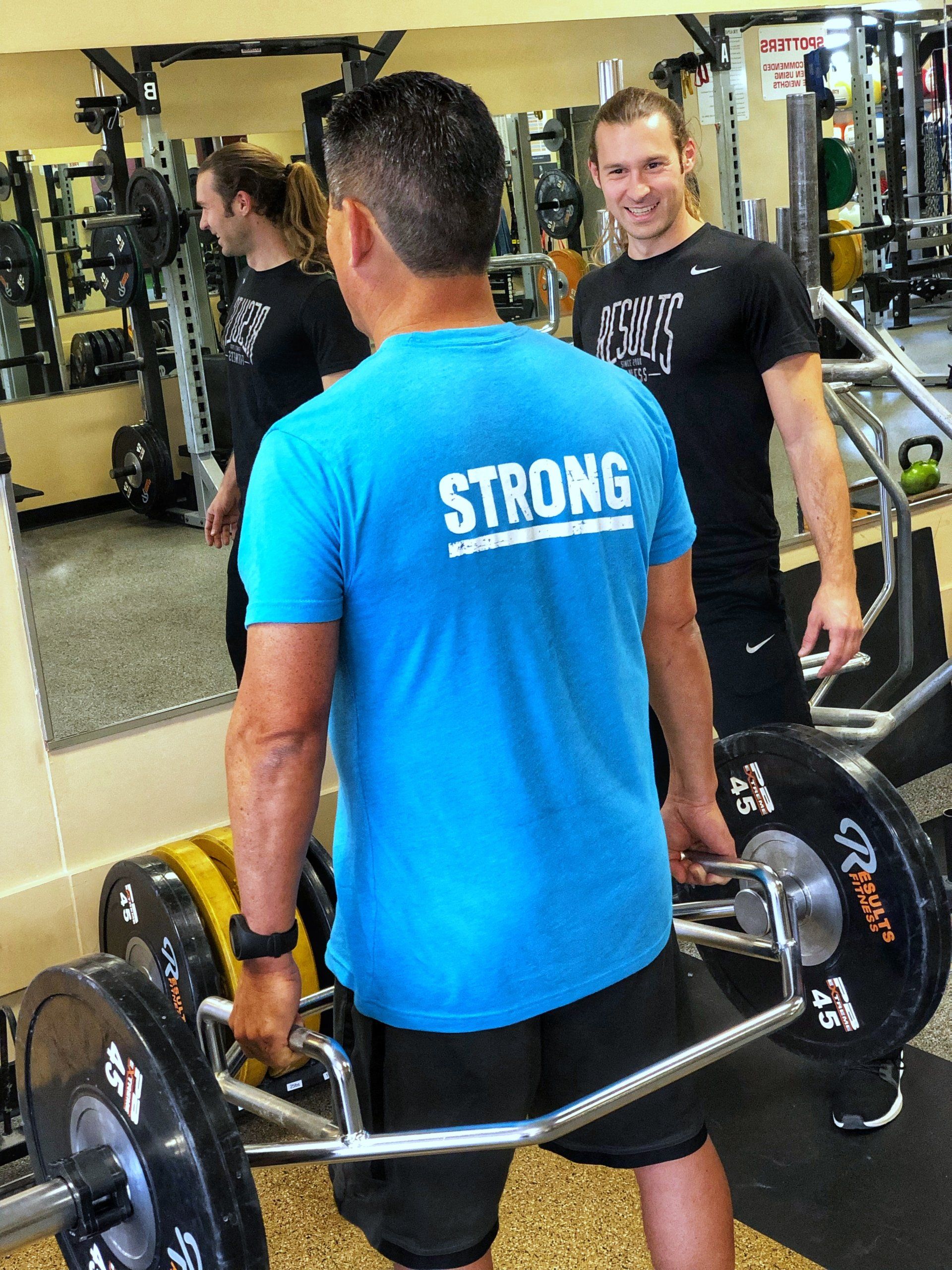 Man lifting weights in gym, another man observing. Man in blue shirt with