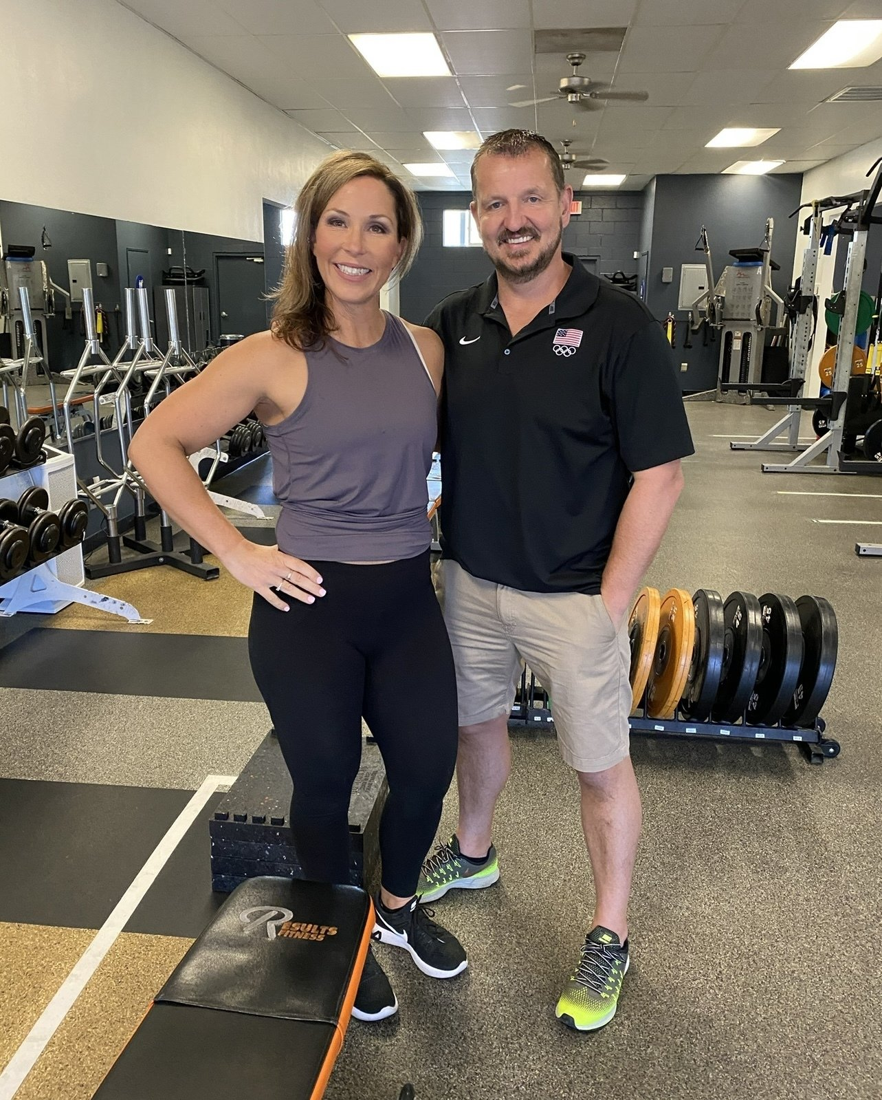 Woman and man in gym setting. Woman in workout clothes, man in polo shirt. They stand together, smiling.