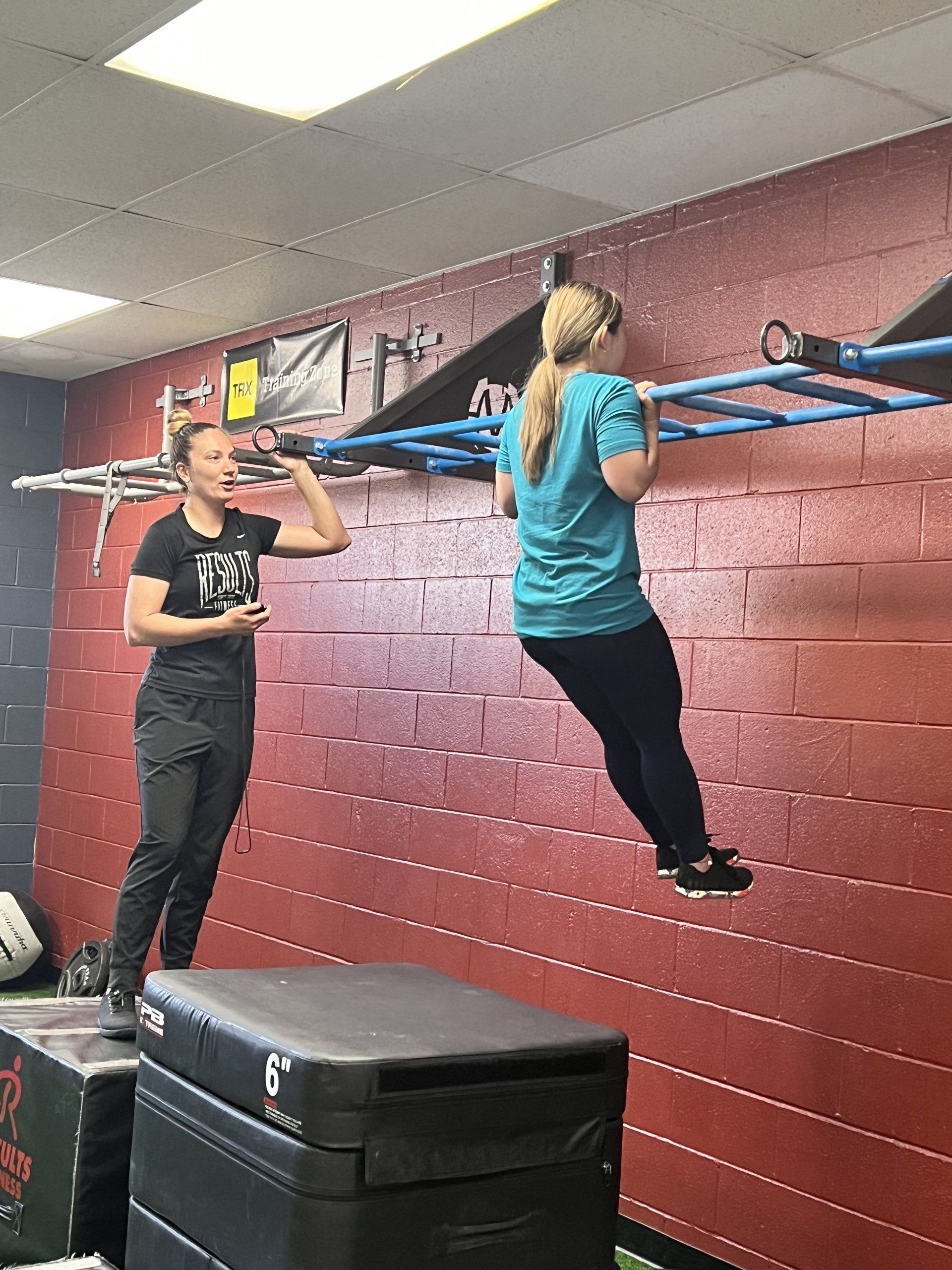 Woman doing pull-up with trainer's guidance. Gym setting with red wall and blue climbing bars.