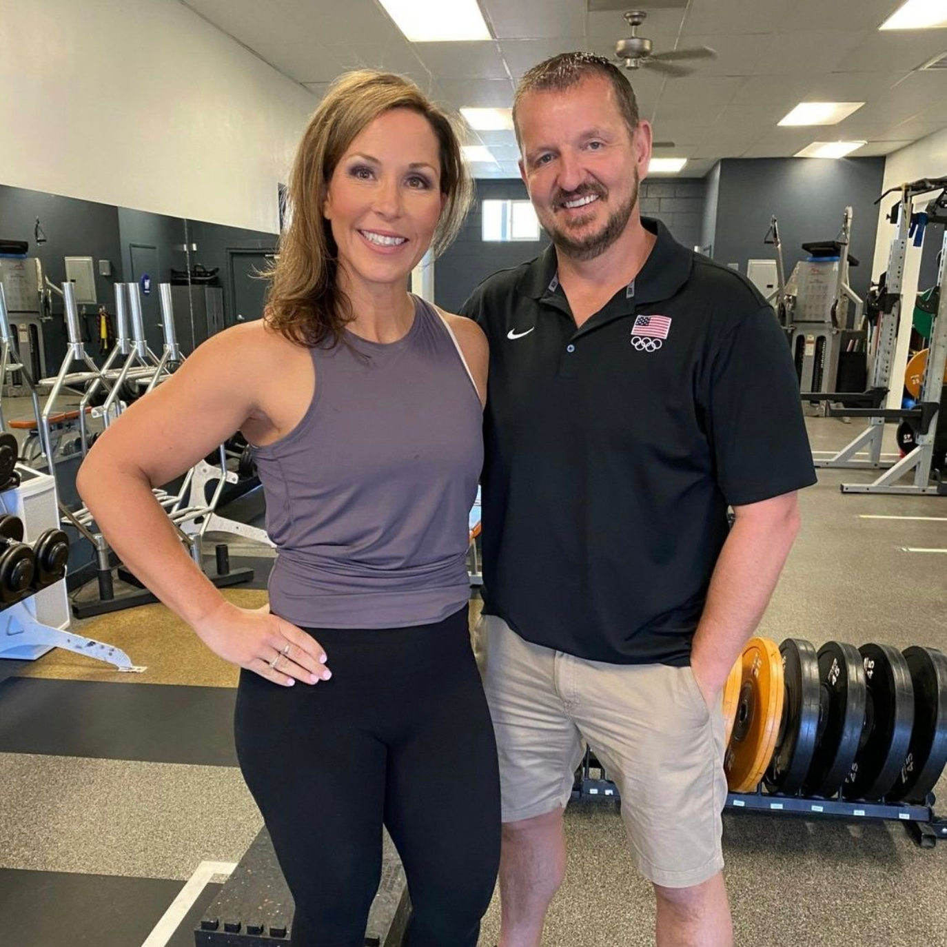 Woman and man smiling in a gym. She wears athletic wear, he wears a polo shirt and khaki shorts. Gym equipment in background.