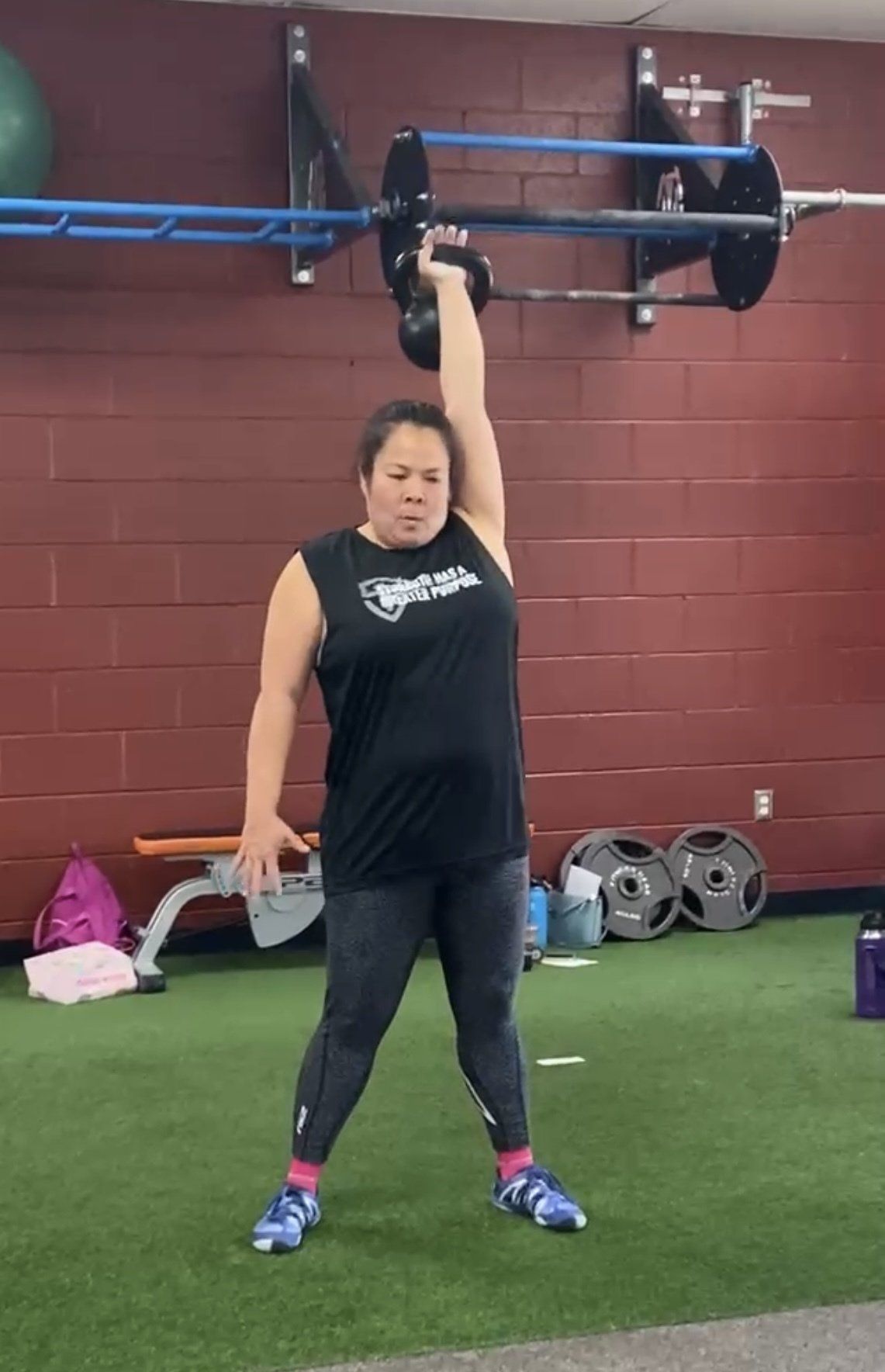 Woman in gym lifting a kettlebell overhead with one arm. She stands with legs apart on green turf.