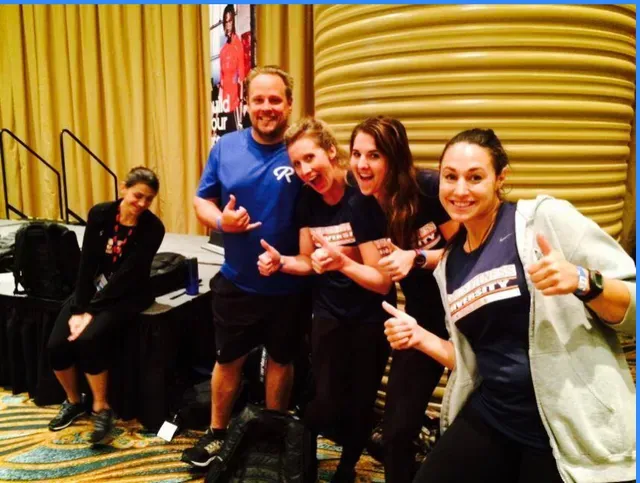 Group of people giving thumbs up indoors; smiling, some wearing matching shirts.