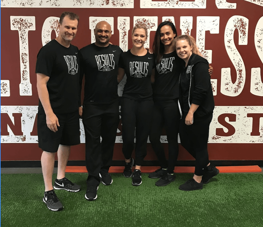 Five people wearing black shirts pose in front of a wall with text. They stand on green turf.