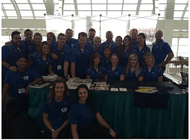 Group of people wearing blue shirts posing behind a table, possibly at an event.