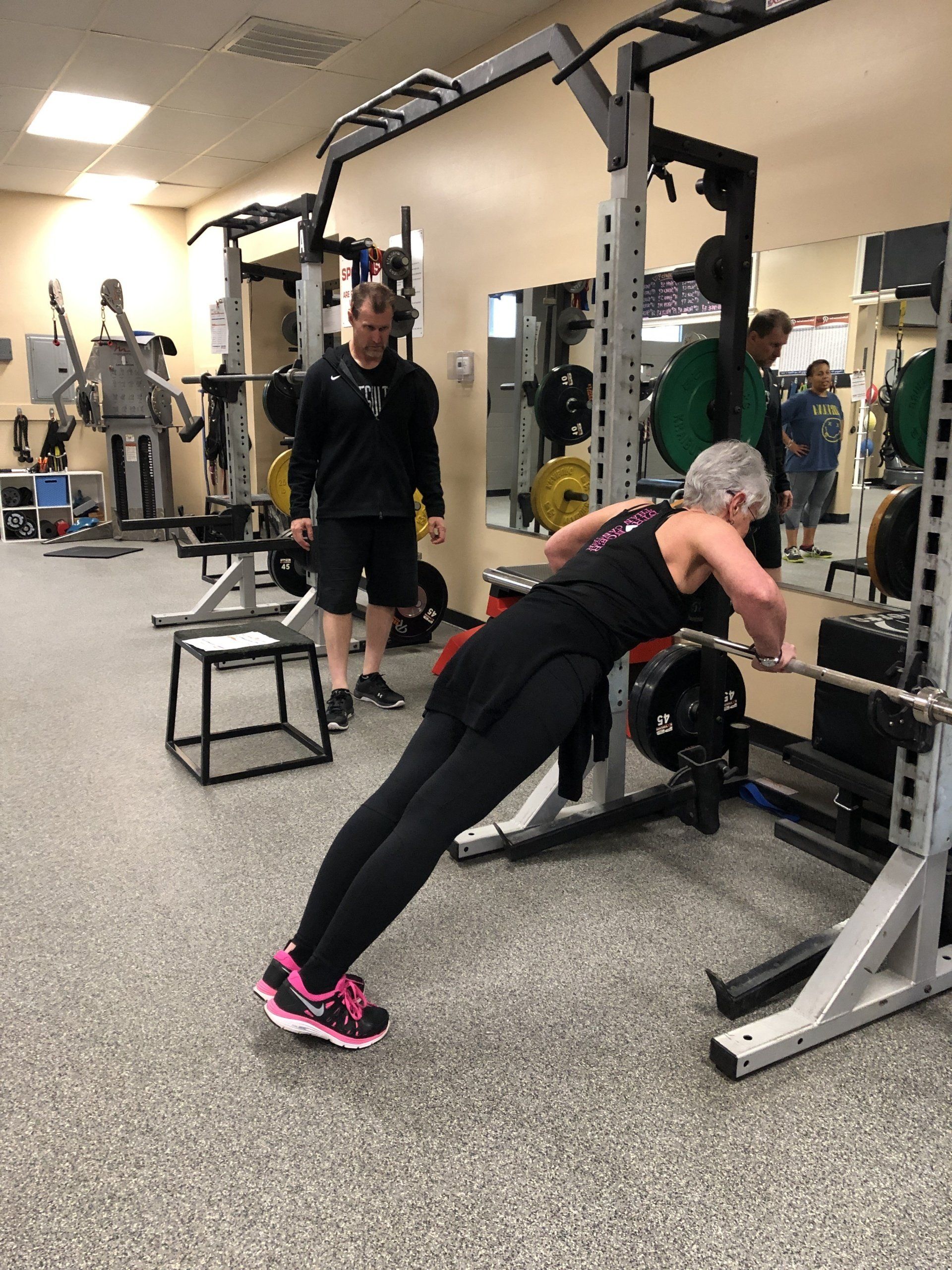 Woman performing a barbell row in a gym, assisted by a coach.