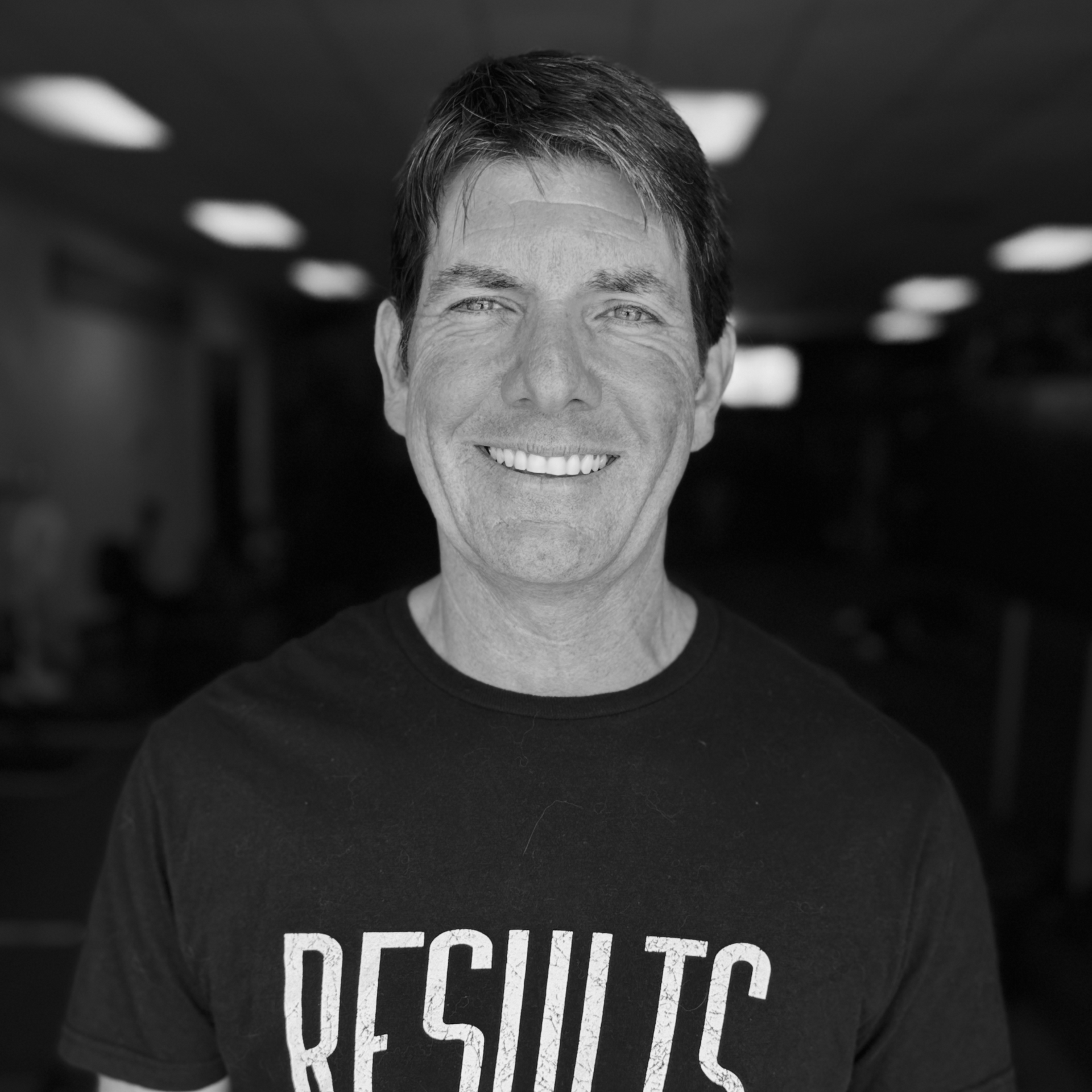 Smiling man in a black shirt with “RESULTS” text, standing in a dim indoor hallway.