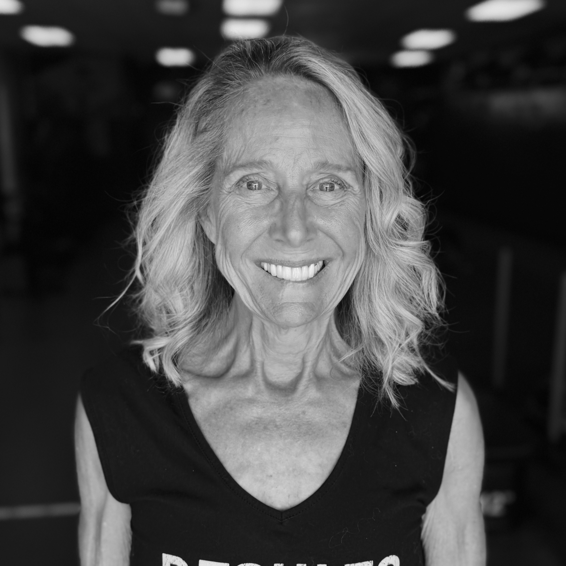 Smiling woman with shoulder-length hair in a dark sleeveless top, standing indoors against a blurred background
